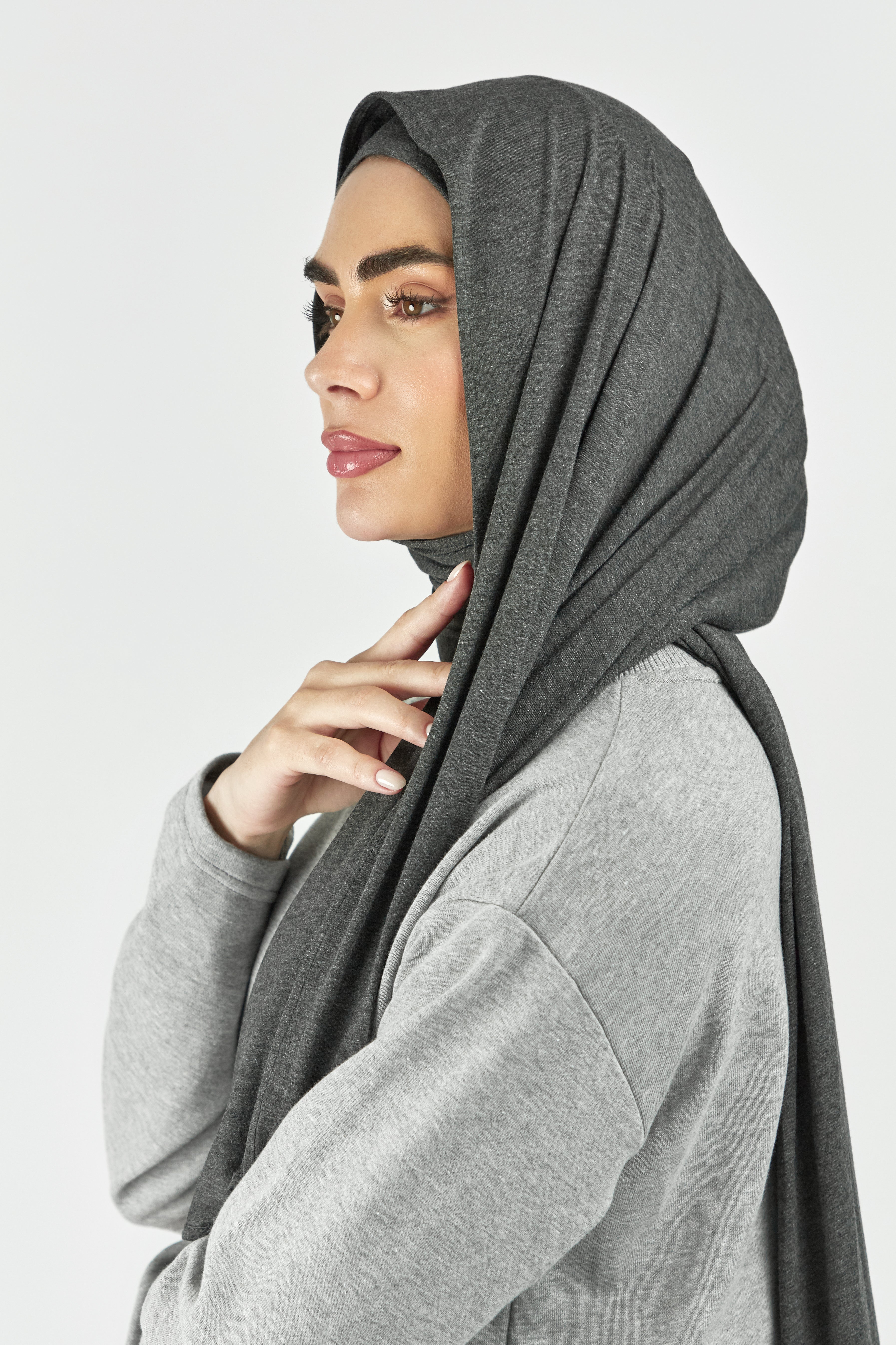 A close-up of a woman wearing one of FARES's matching jersey hijab sets. The model is turned to the left. Colour description: dark heather grey. This set includes a modal jersey hijab and matching undercap.