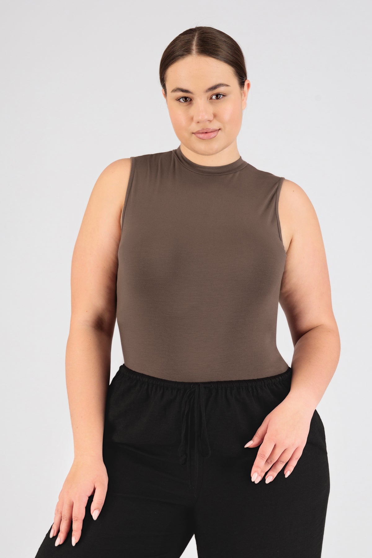 The front of a model standing in FARES' sleeveless layer with her hands on her pants. Colour description: Ash Brown.