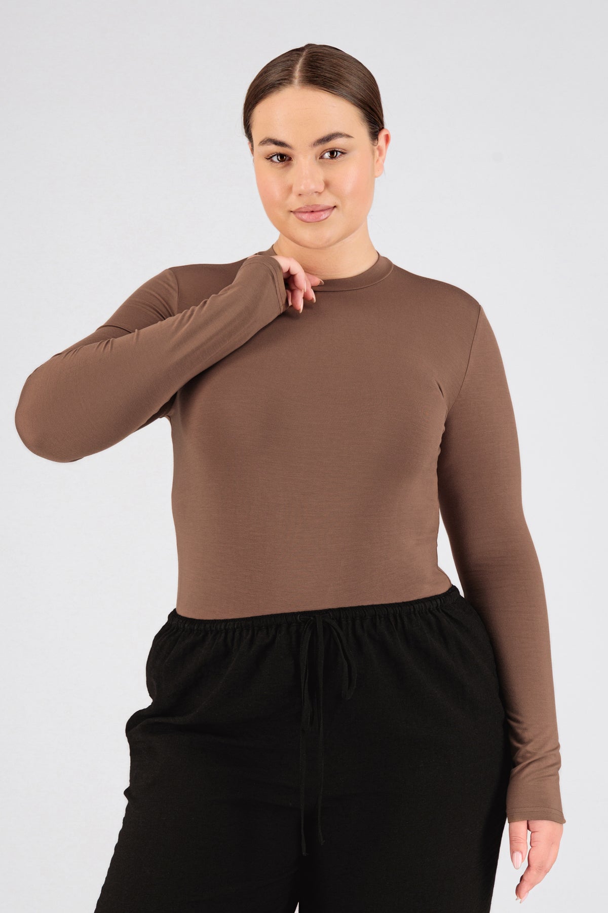 A model is wearing FARES' fitted long sleeve. She is standing in this base layer clothing with perfect fit sleeves with her hand on her neck. Colour description: Bronze.