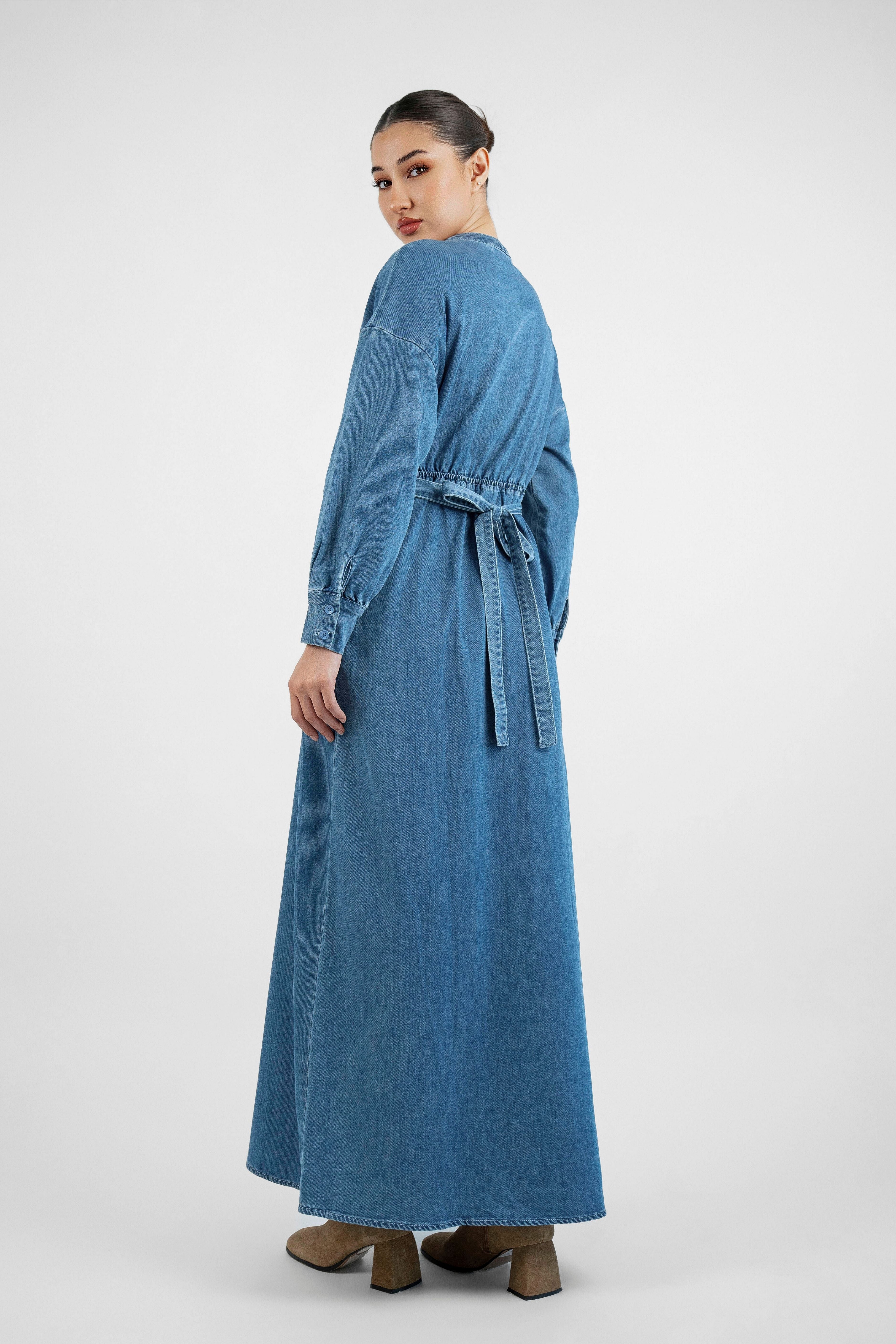 A girl is standing with her back looking over her left shoulder in FARES' denim dress. Colour description: Blue.