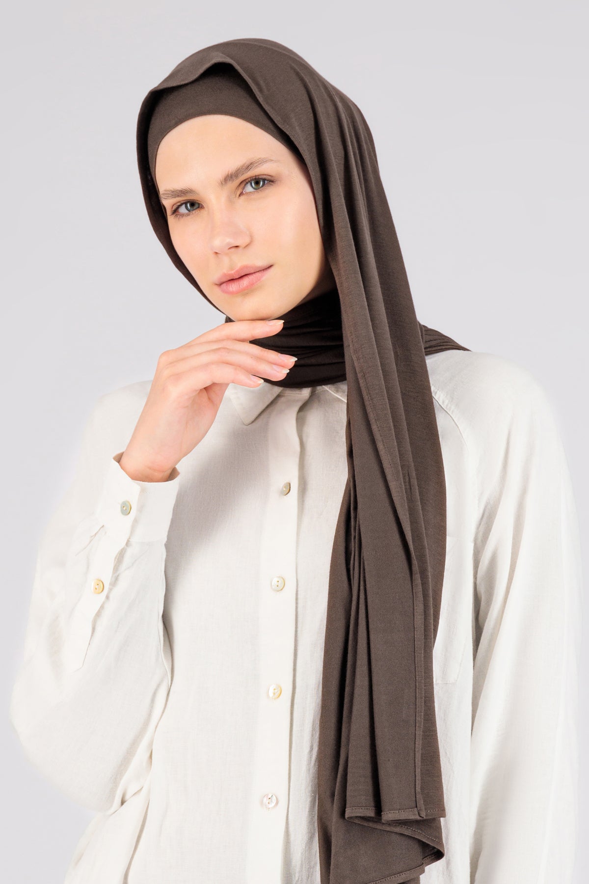 A woman wearing one of FARES's matching jersey hijab sets. Colour description: Cool Dark Brown. This set includes a modal jersey hijab and matching undercap.