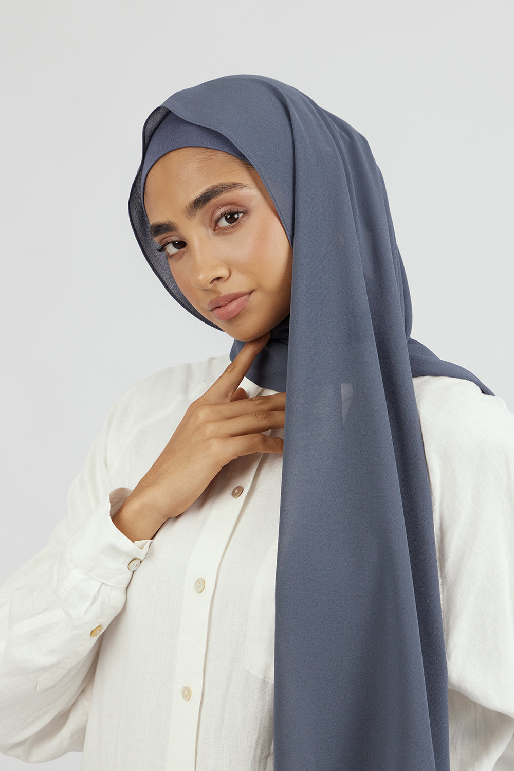 The front of a woman touching the matching chiffon hijab set she wears. Colour description: denim blue.