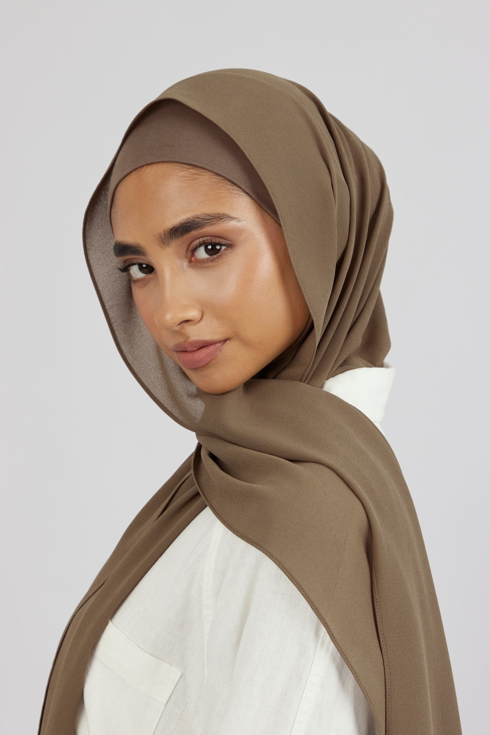 A close-up of a female model wearing a matching chiffon hijab set. The model is standing side ways and looking over her left shoulder. Colour description: medium beige-brown.