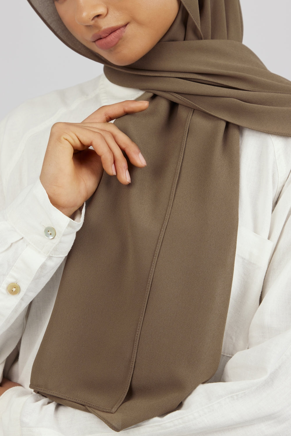 A close-up of a female model touching the matching chiffon hijab set she wears. Colour description: medium beige-brown.