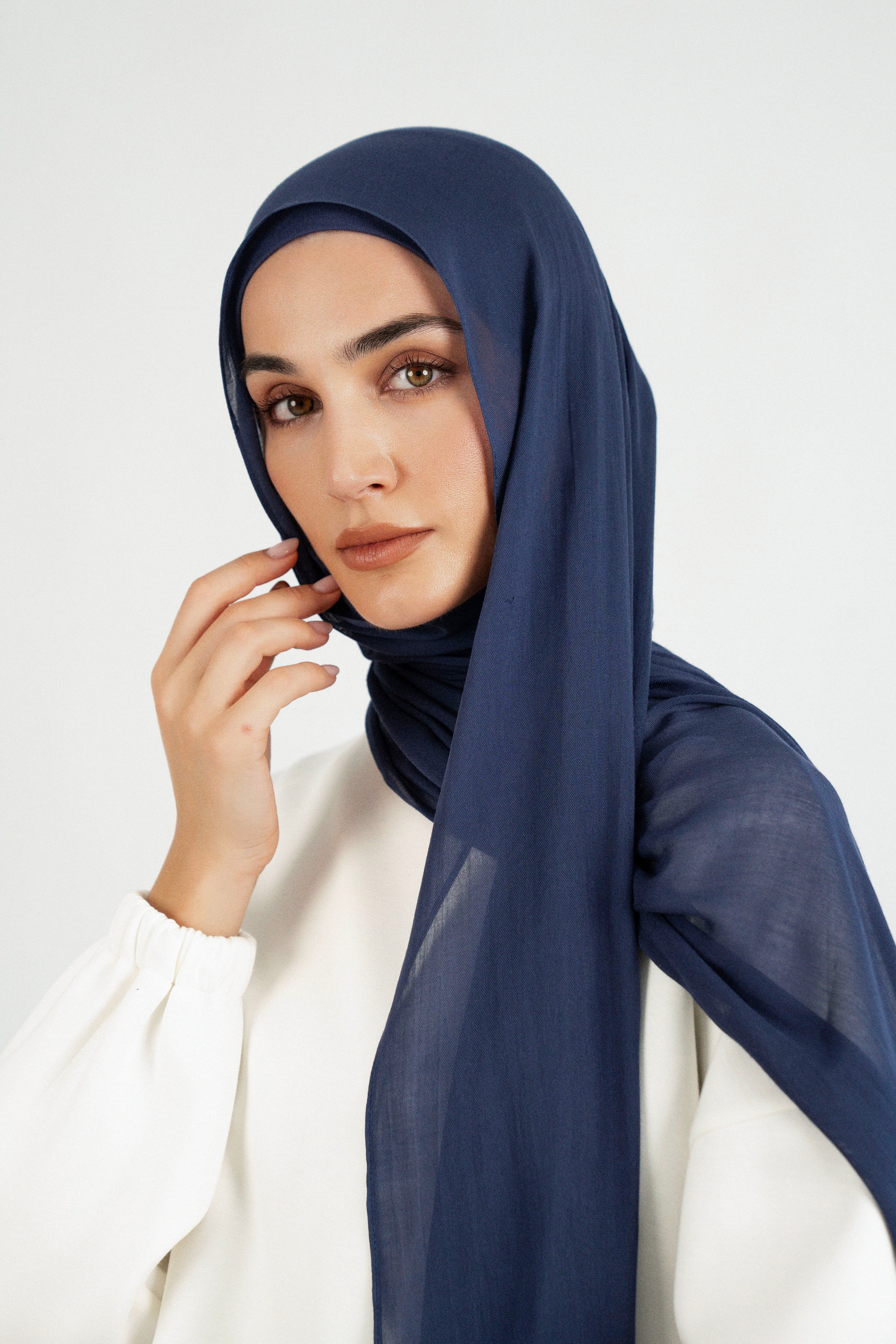 Colour: Navy. A close up of a woman wearing a Matching Butter Modal Hijab Set and touching her face.