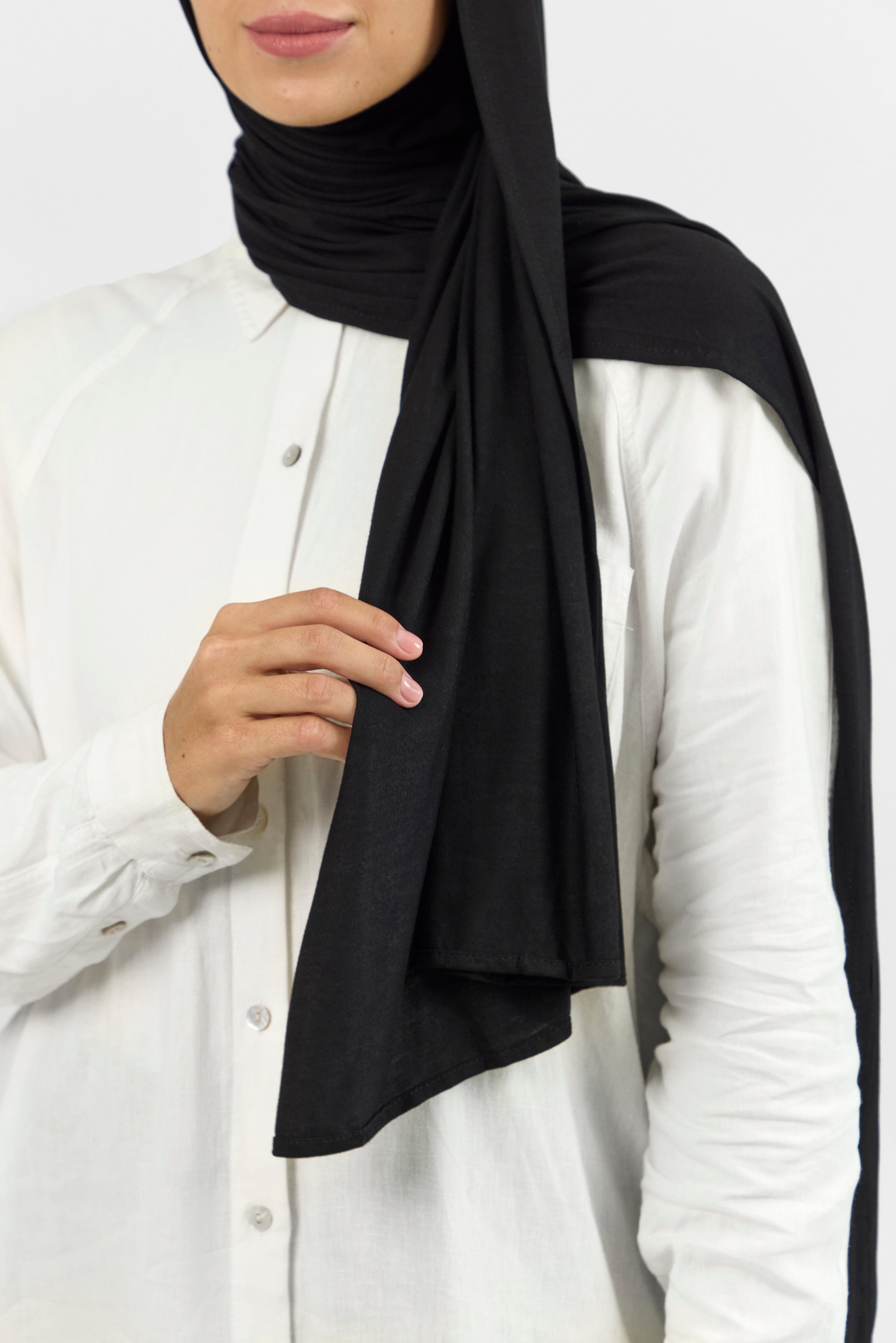 A close-up of the front of a female model touching the matching jersey hijab set she wears. Colour description: black.