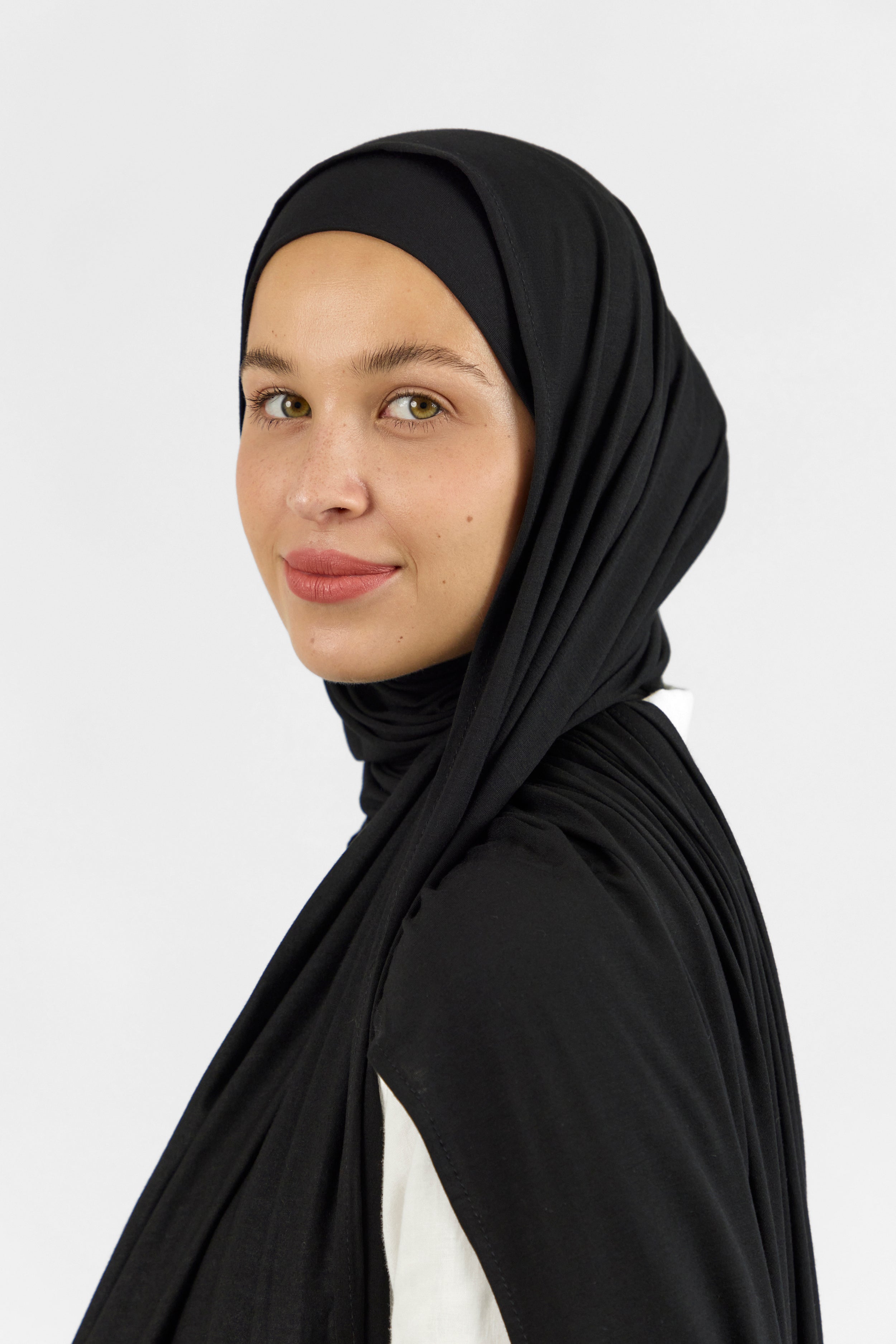 A female model wearing a matching jersey hijab set. Colour description: black. The model is standing side ways and looking over her left shoulder. This set includes a modal jersey hijab and matching undercap.
