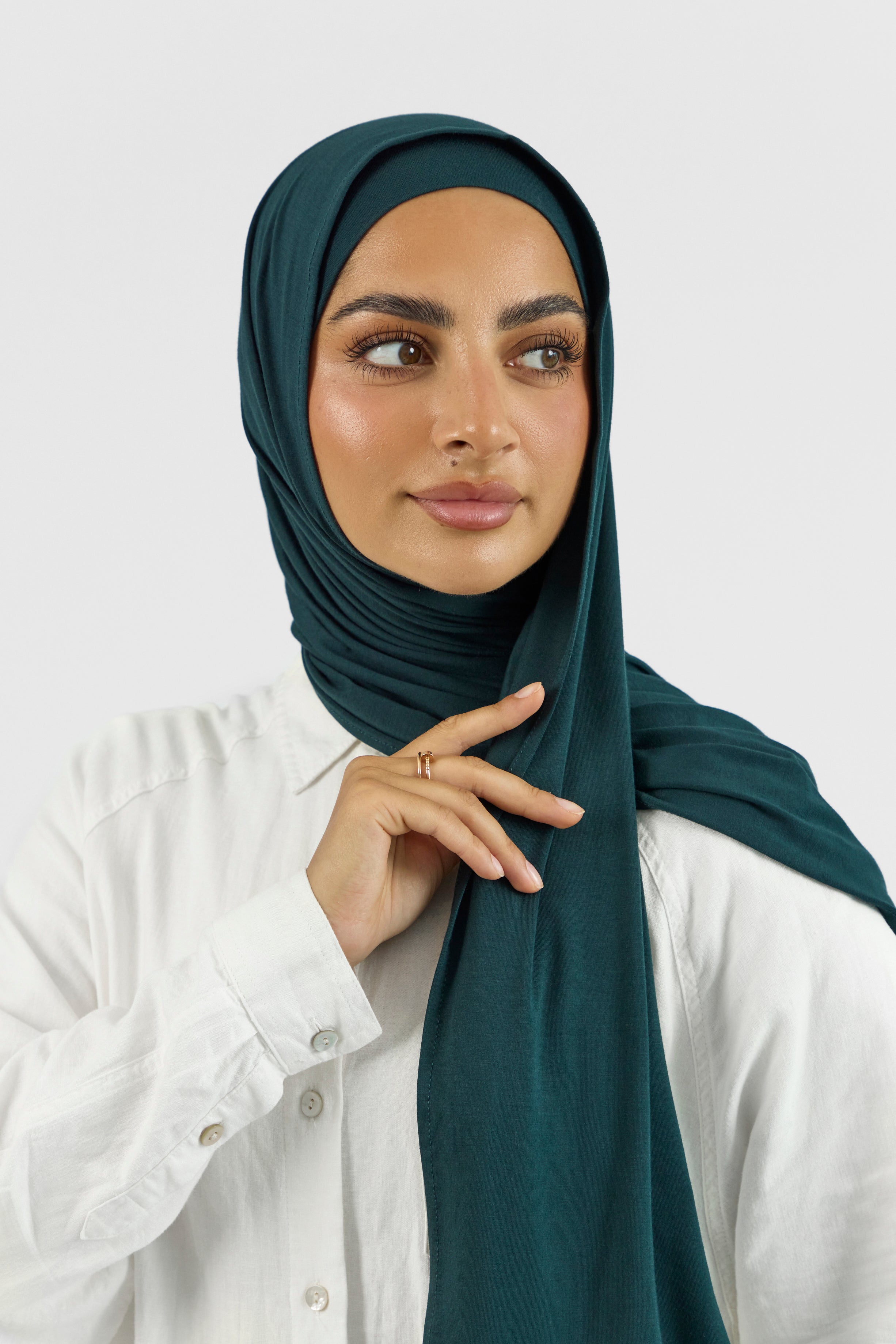A close-up of a female model touching the matching jersey hijab set she wears. Colour description: dark teal.
