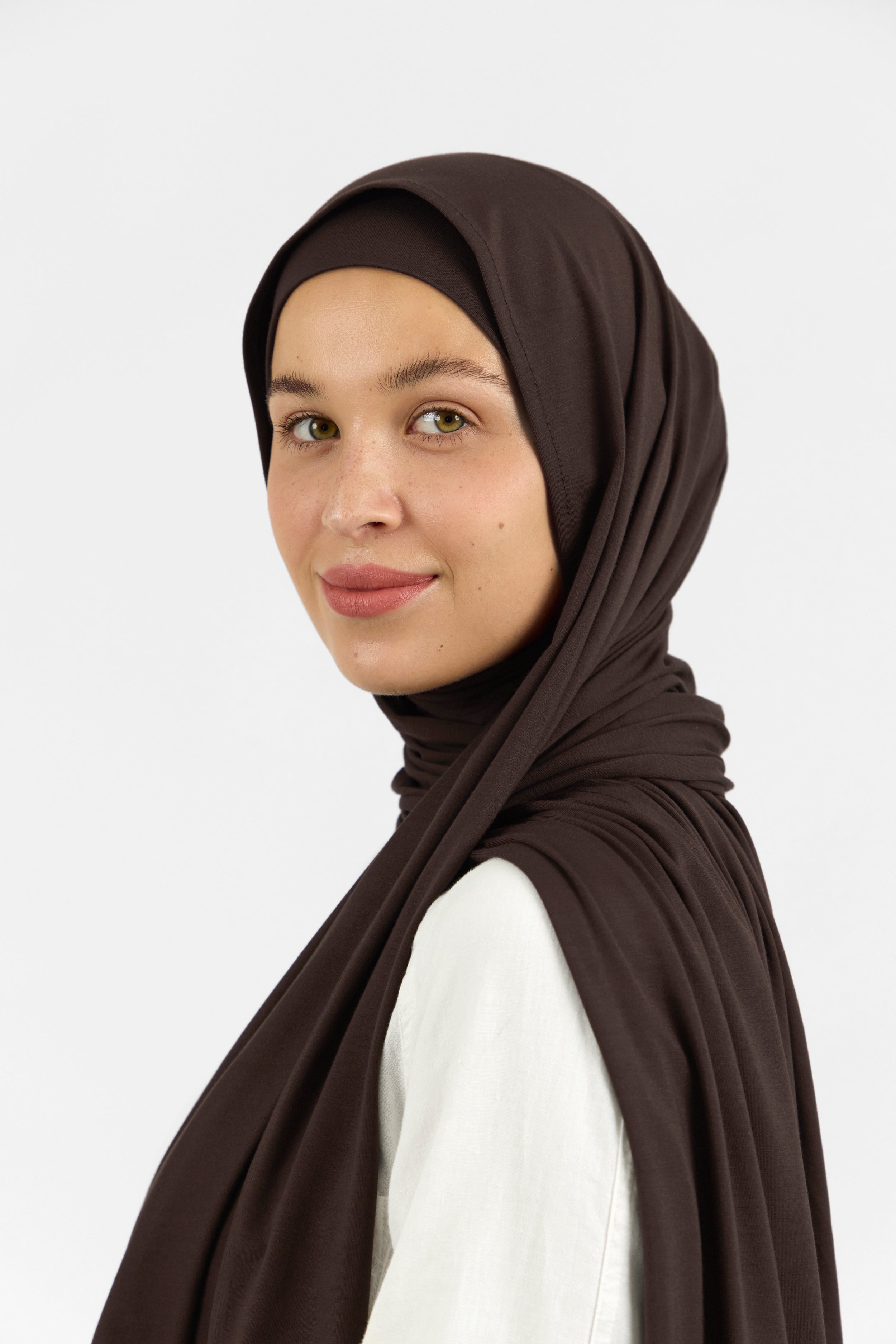 A female model wearing a matching jersey hijab set. The model is standing side ways and looking over her left shoulder. Colour description: dark brown.
