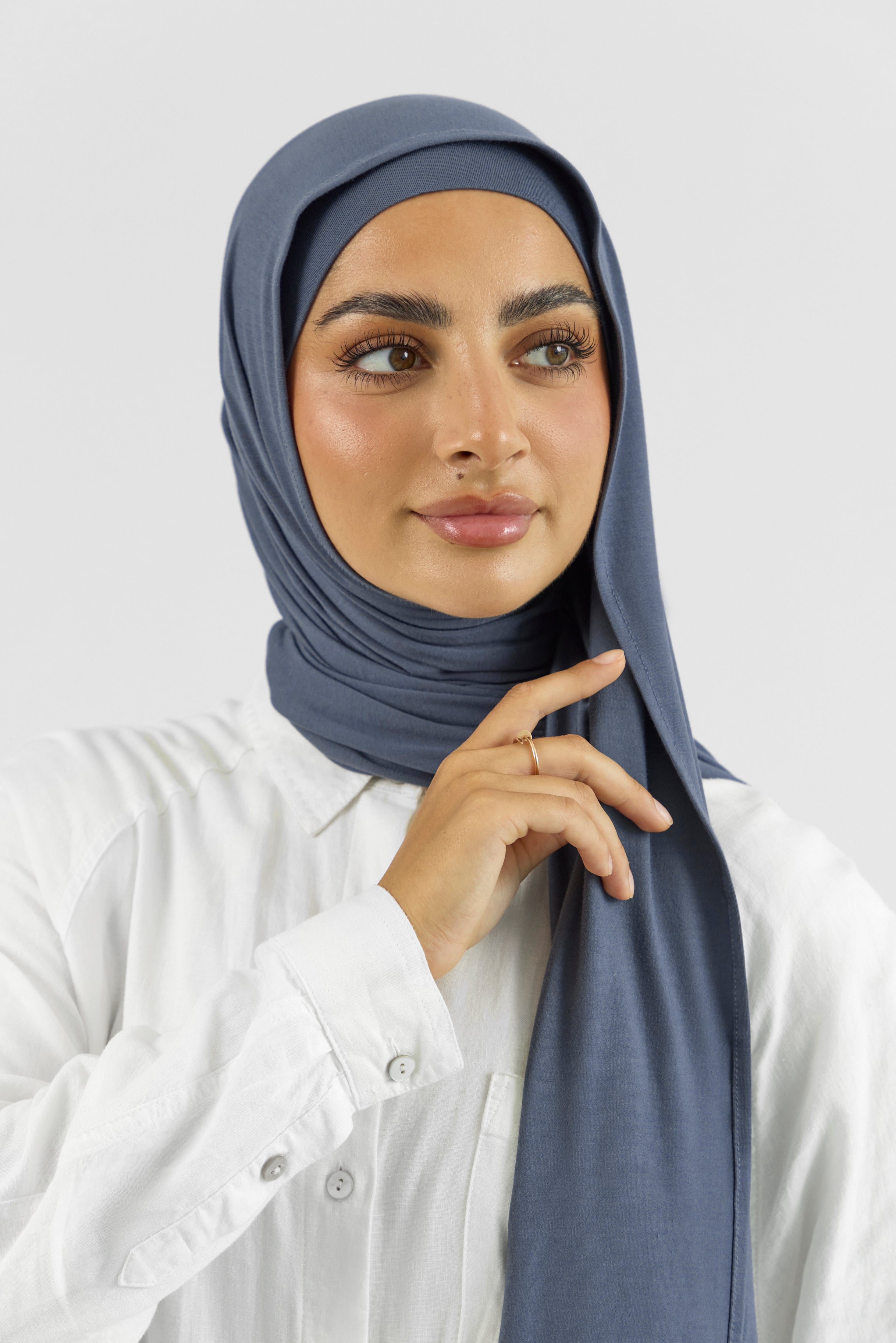 The front of a female model touching the matching jersey hijab set she wears. This set includes a modal jersey hijab and matching undercap. Colour description: denim blue.