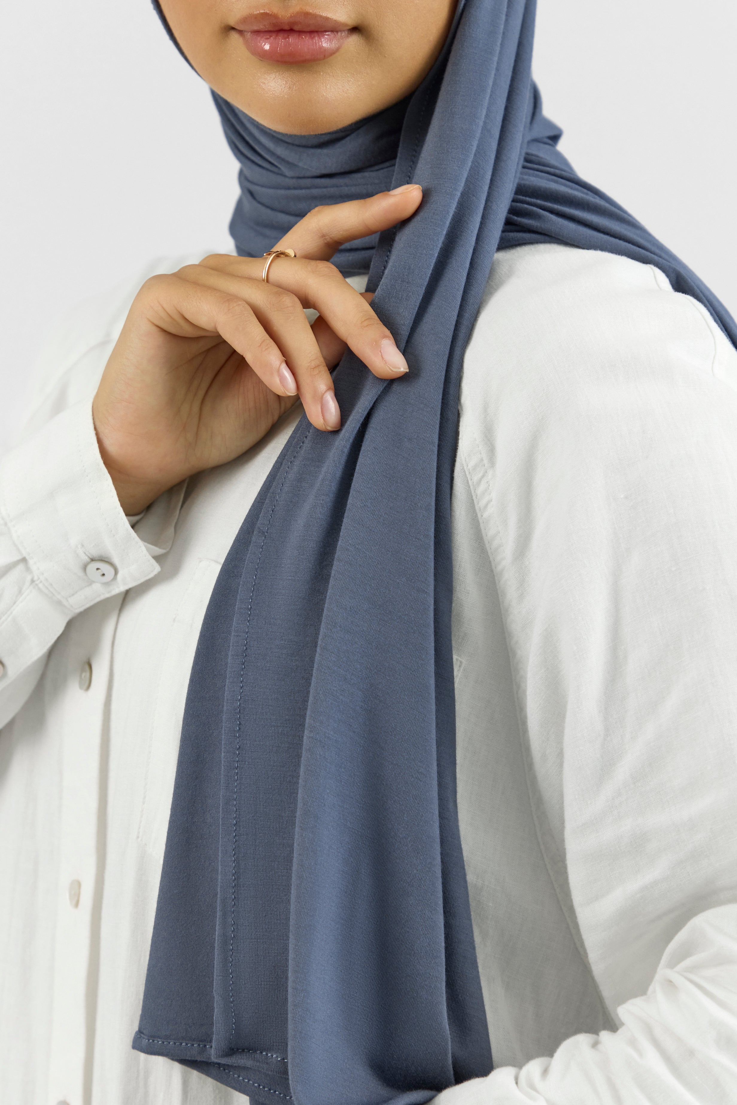 A close up of the front of a female model touching the matching jersey hijab set she wears. This set includes a modal jersey hijab and matching undercap. Colour description: denim blue.