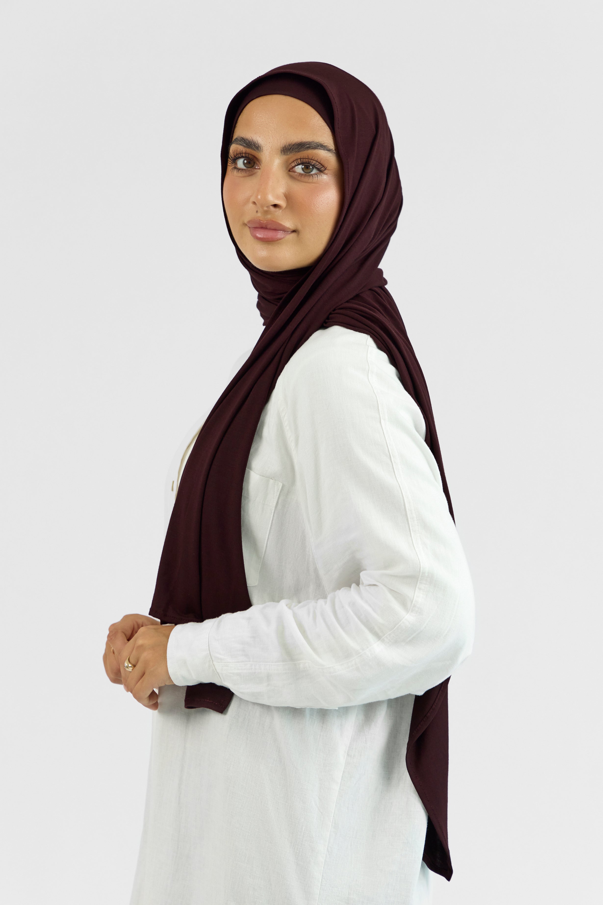 A female model wearing a matching jersey hijab set. The model is standing side ways and looking over her left shoulder. Colour description: deep plum.