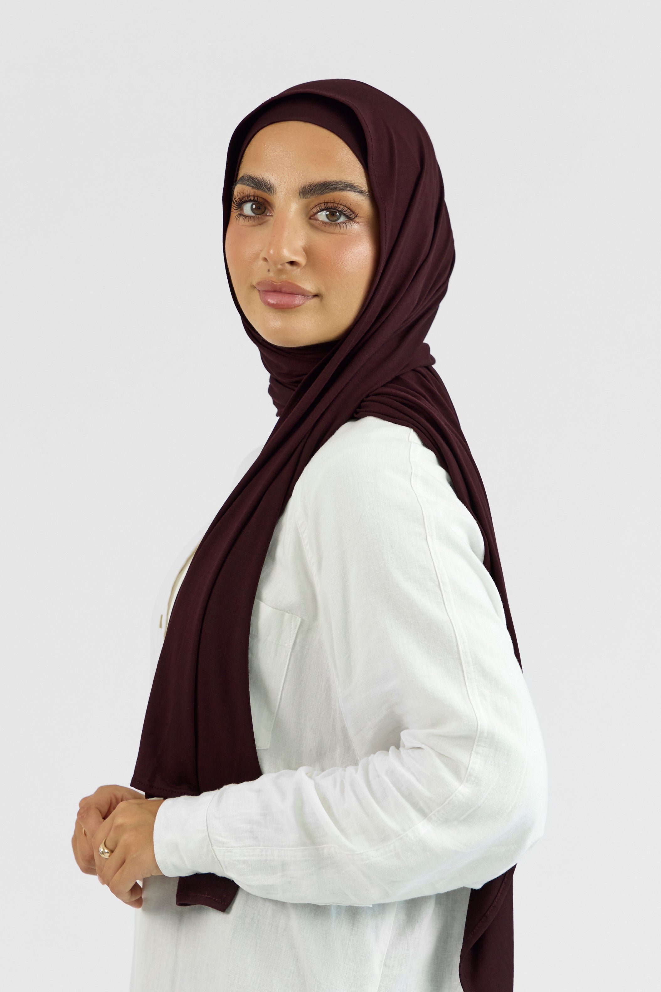 A female model wearing a matching jersey hijab set. The model is standing side ways and looking over her left shoulder. Colour description: deep plum.