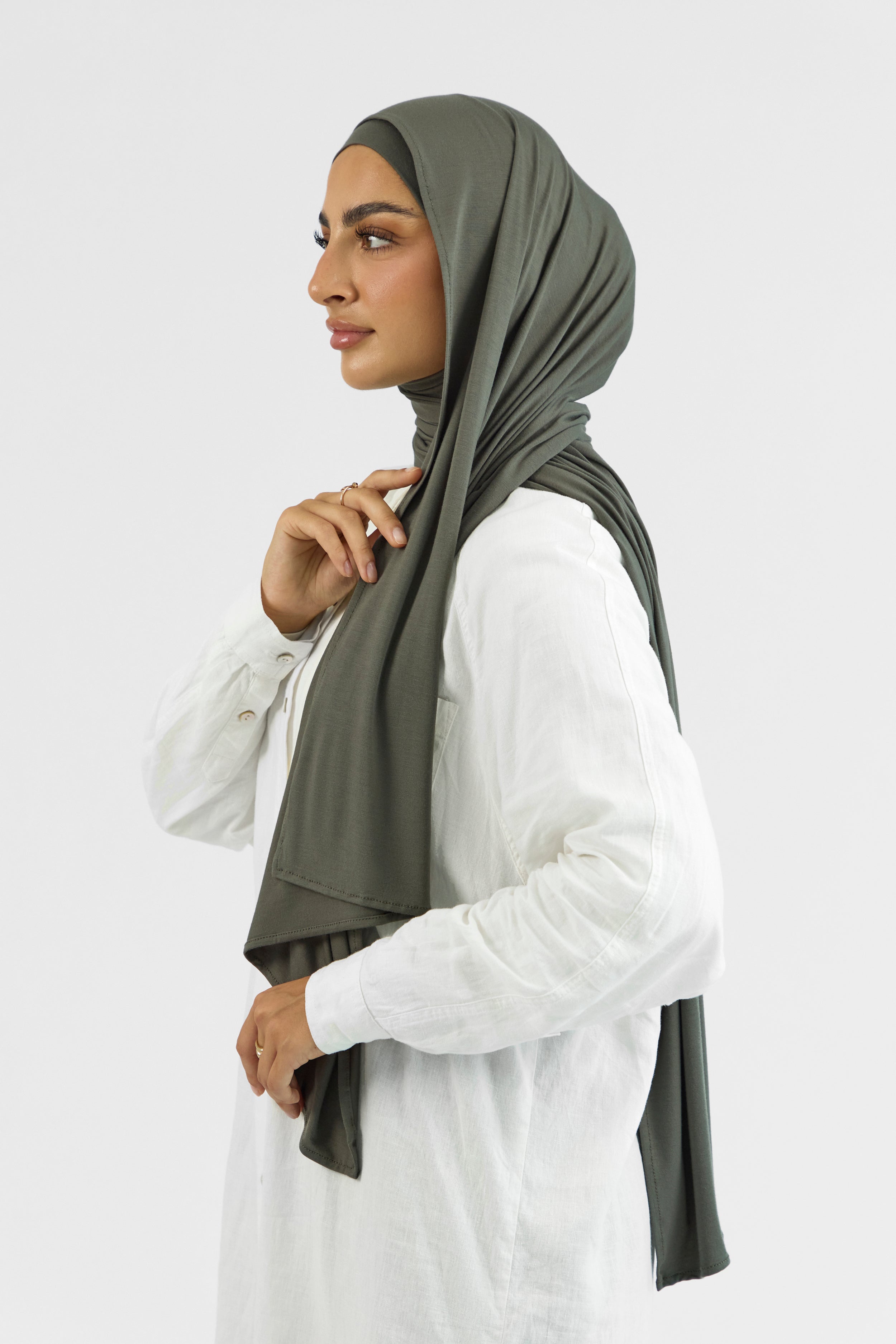 A woman wearing one of FARES's matching jersey hijab sets. The model is turned to the left. Colour description: muted green. This set includes a modal jersey hijab and matching undercap.