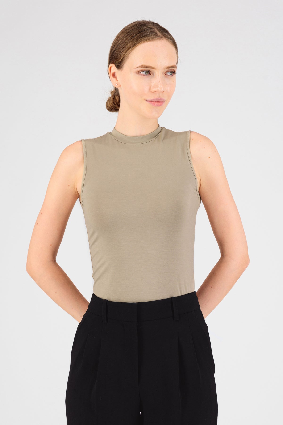 A model is wearing FARES' sleeveless layer. Colour description: Sand. She is standing in the sleeveless top with her hands behind her back..