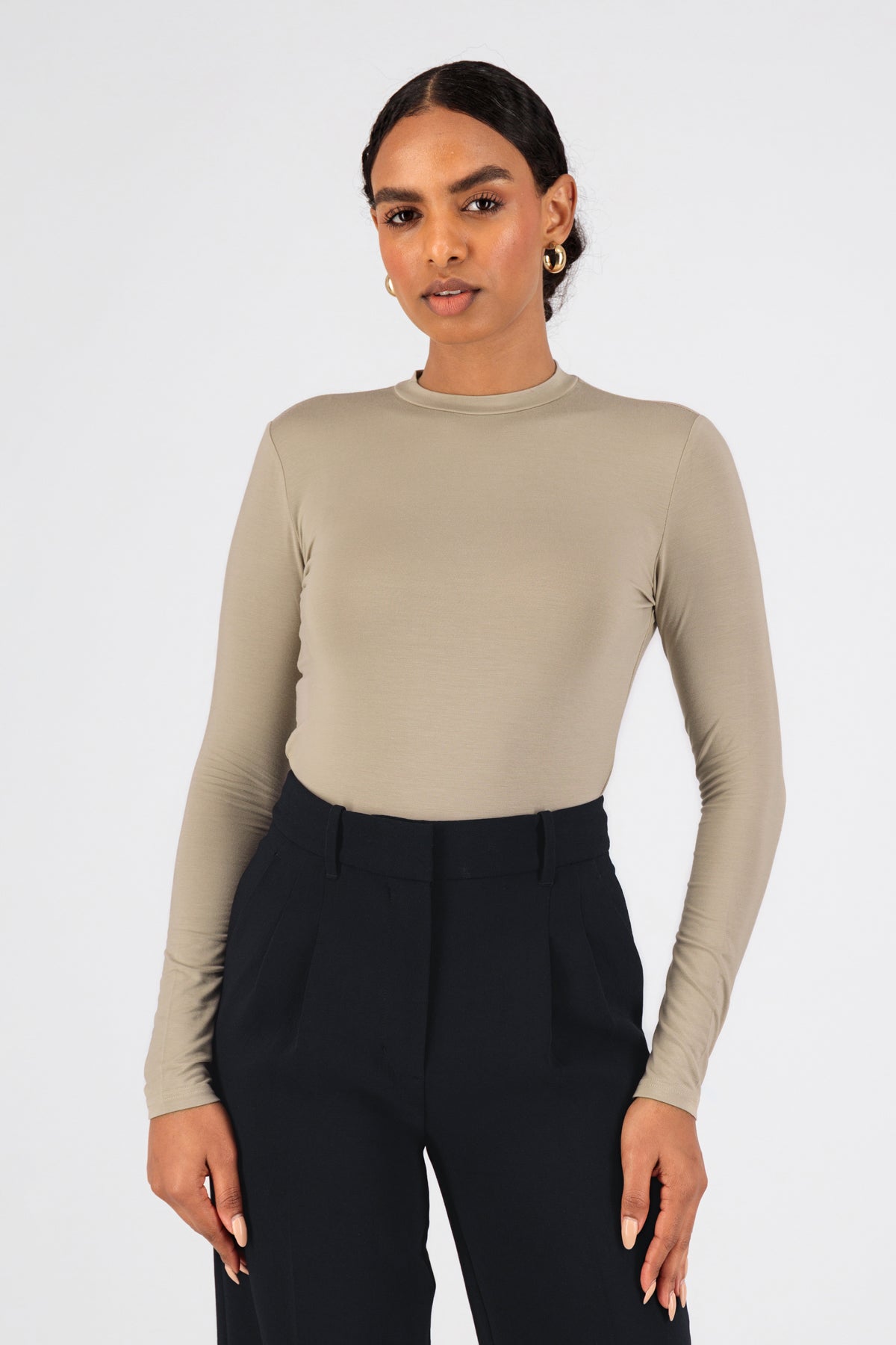 A model is wearing FARES' long sleeve layer. Colour description: Sand. She is standing with her front to the camera in a fitted long sleeve base layer.