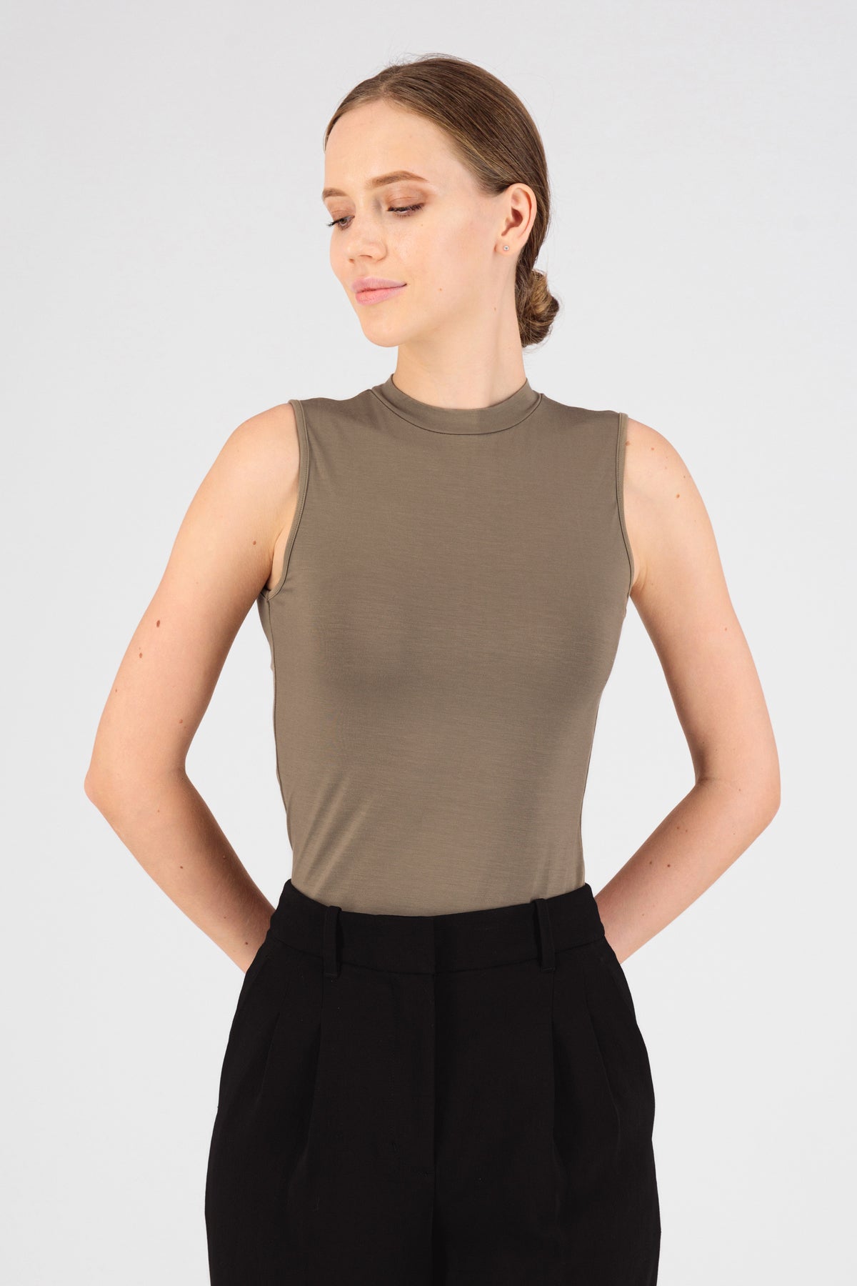 A girl is wearing FARES' sleeveless layer. Colour description: Taupe. She is standing in the sleeveless top with her hands behind her back..