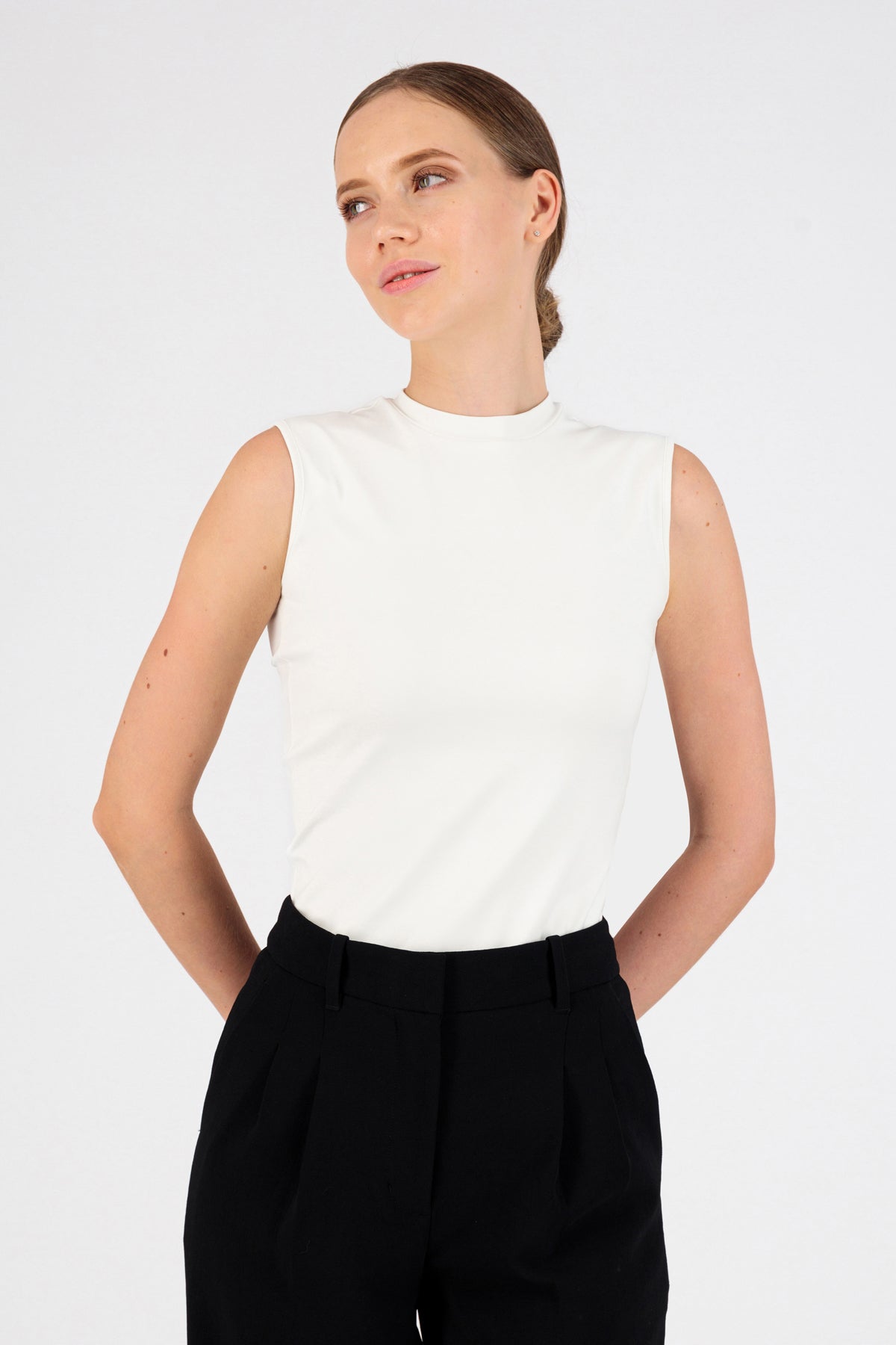 A girl is wearing FARES' sleeveless layer. Colour description: White. She is standing in the sleeveless top with her hands behind her back..