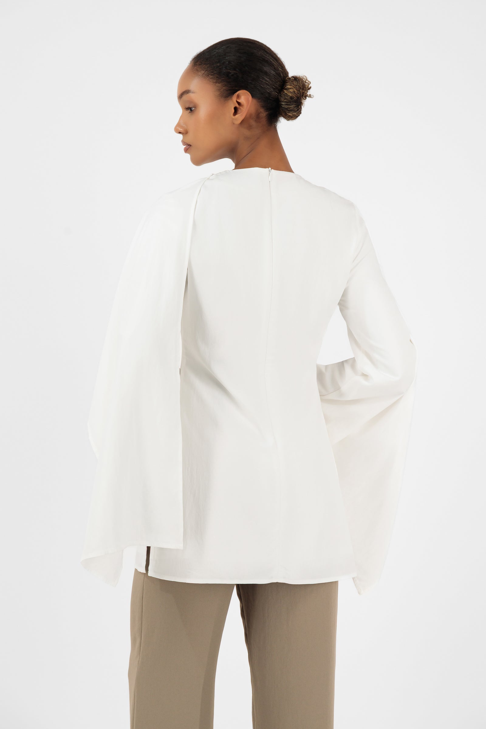 Back view of a woman wearing the white FARES tunic clothing design showcasing the soft cape overlay and relaxed fit.