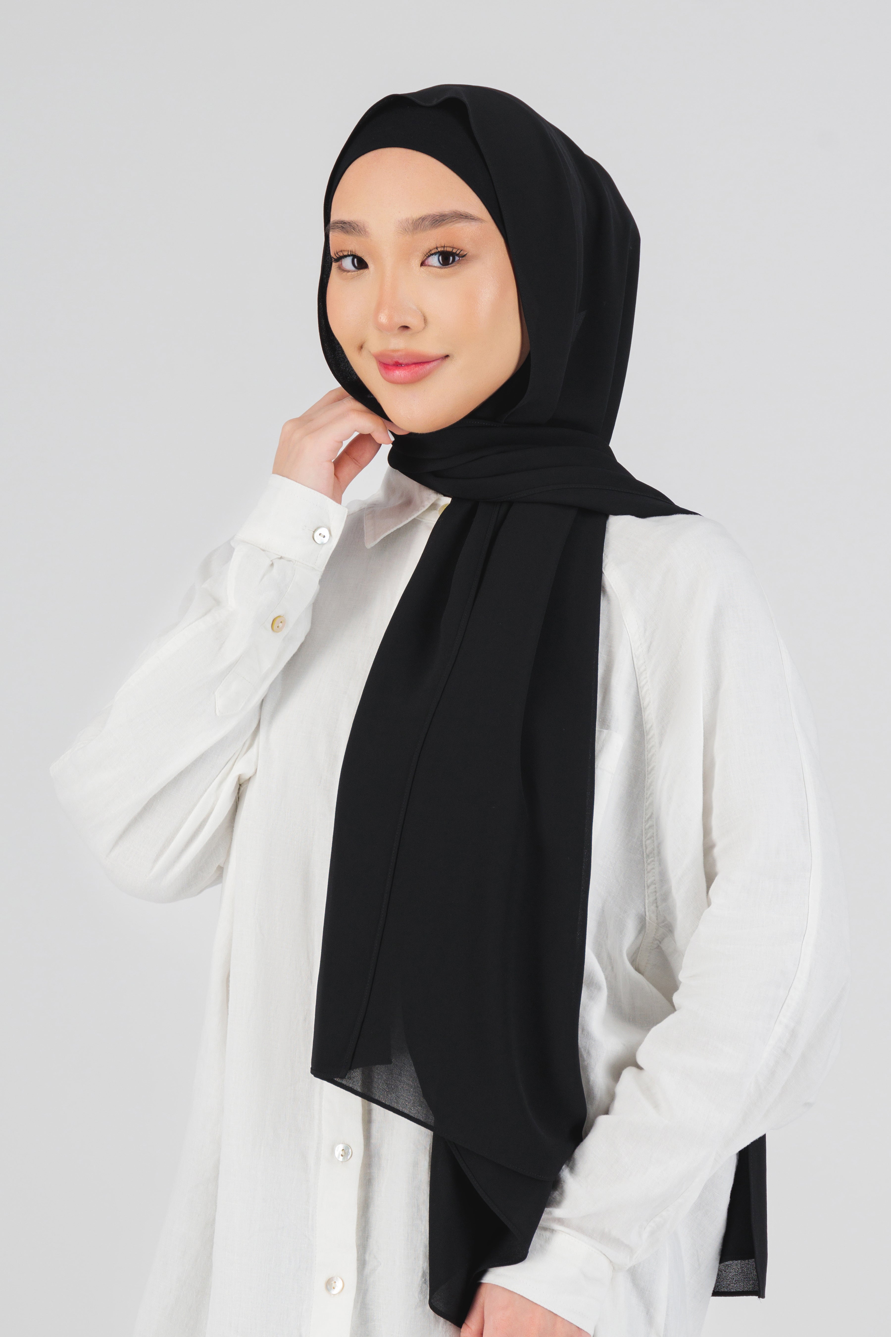 A female model touching the matching chiffon hijab set she wears. The model is turned to the left. This hijab fashion wear includes a high quality chiffon hijab and a breathable matching modal jersey undercap. Colour description: black.