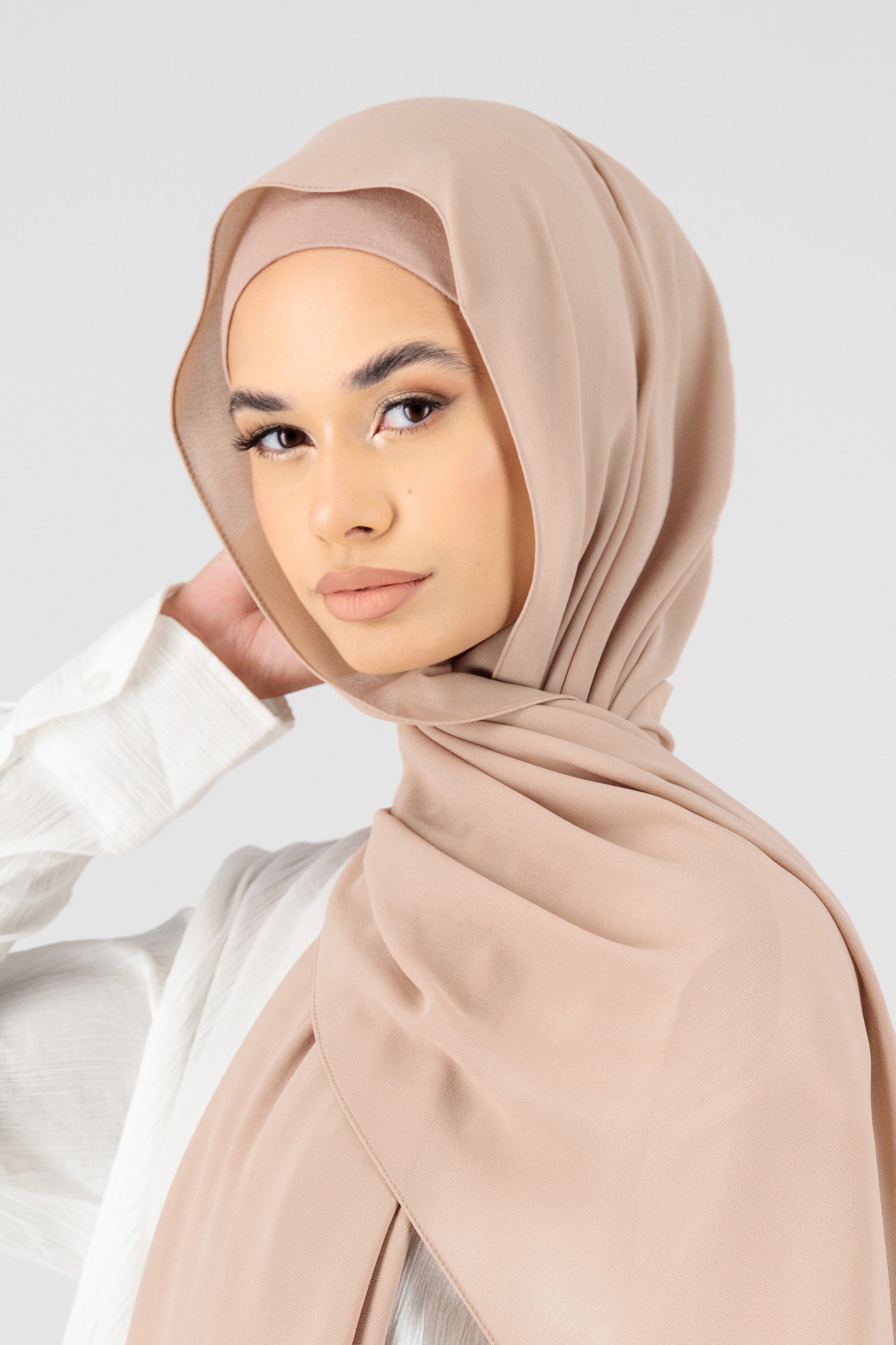 A woman touching the matching chiffon hijab set she wears. This hijab fashion wear includes a high quality chiffon hijab and a breathable matching modal jersey undercap. Colour description: muted pink beige.