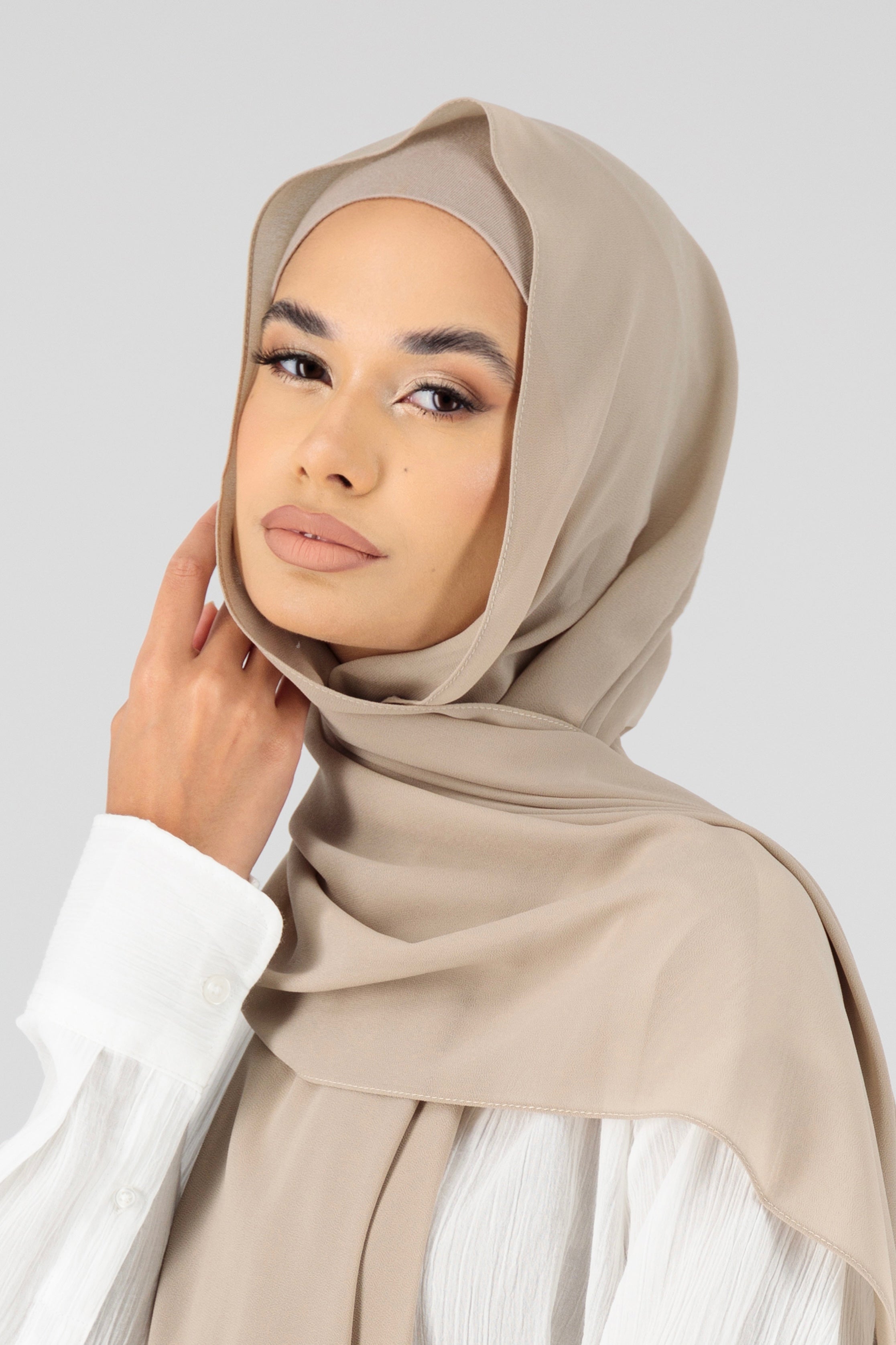 A woman touching the matching chiffon hijab set she wears. This hijab fashion wear includes a high quality chiffon hijab and a breathable matching modal jersey undercap. Colour description: light beige.