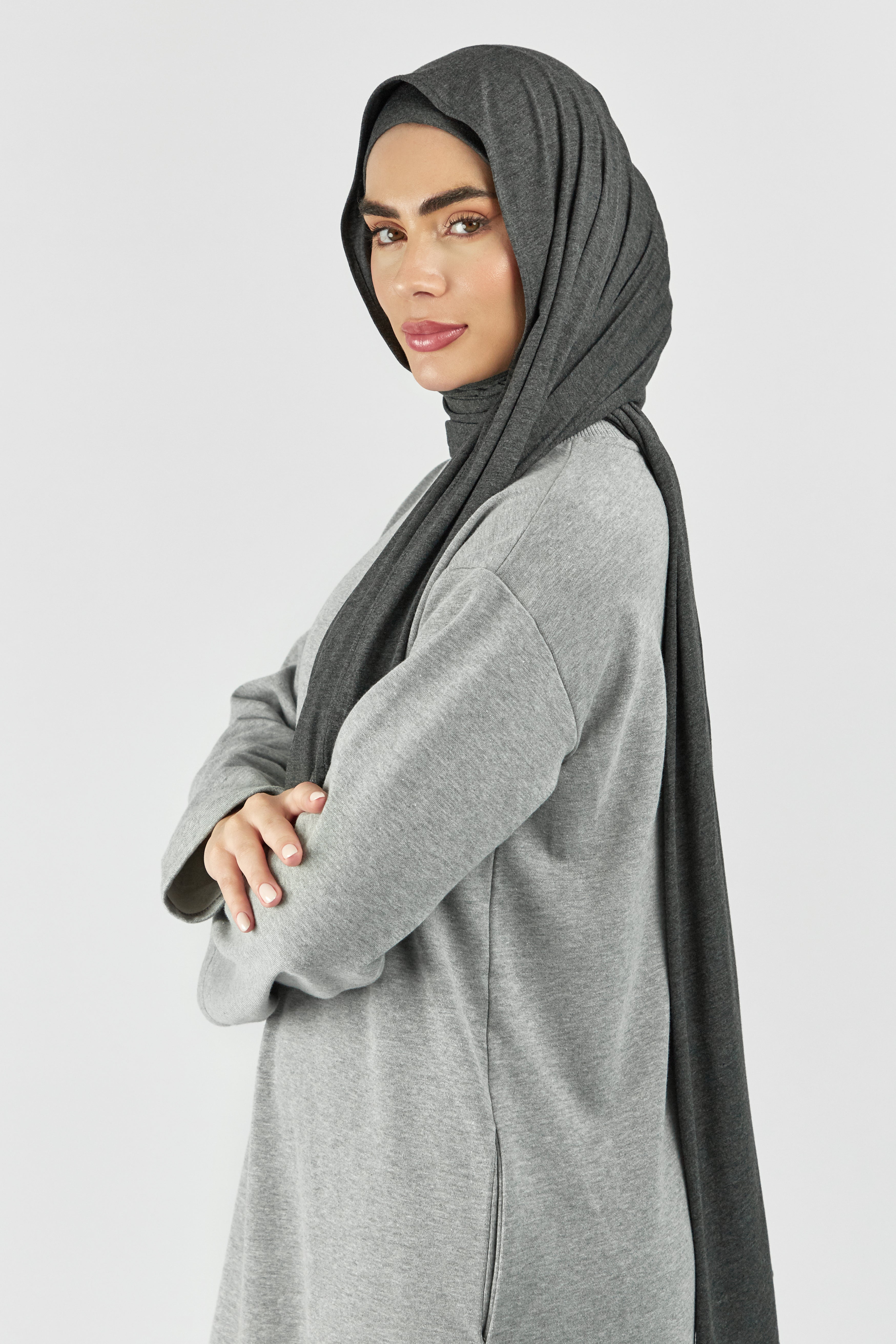 A woman wearing one of FARES's matching jersey hijab sets. The model is turned to the left. Colour description: dark heather grey. This set includes a modal jersey hijab and matching undercap.
