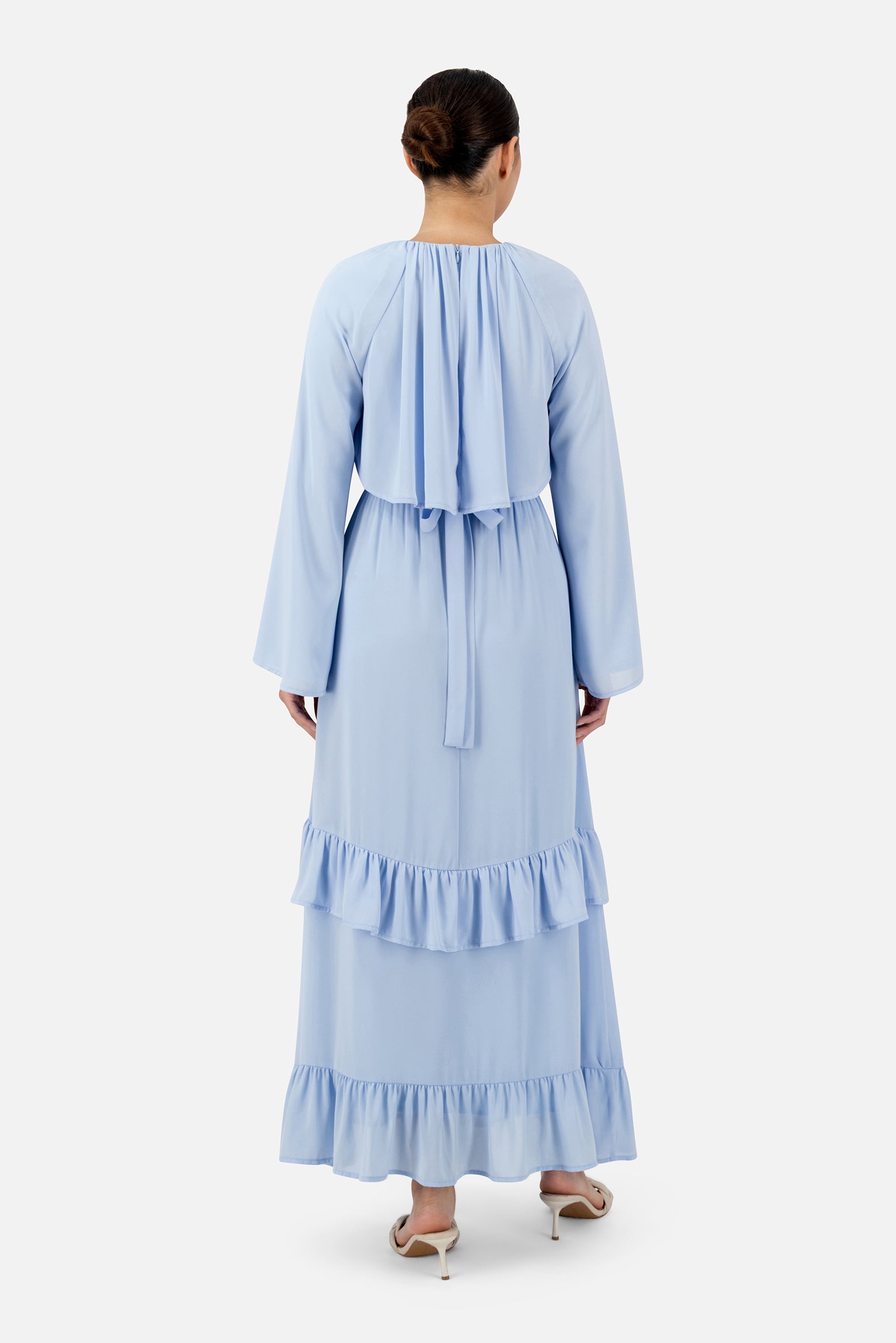 Back view of a woman wearing the powder blue FARES long sleeve maxi dress showcasing the layered back overlay and graceful movement.