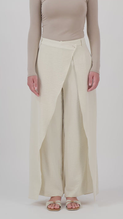 A woman walks in the FARES Skirt Overlay Trousers in Oats, showcasing the movement of the skirt overlay and flowing wide-leg trousers.